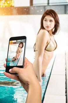 Closely image of female hands holding mobile phone with photo camera mode on the screen. Cropped image of beautiful long hair female model posing by the pool