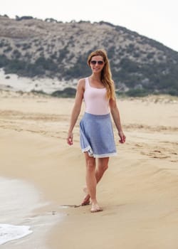 Young woman in blue skirt, t shirt and sunglasses walking on beach, smiling, overcast weather, hill behind her.