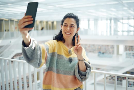 Selfie, phone and peace with a woman in a library for research while posing for a social media photograph. Mobile, communication and hand gesture with a female student taking a picture in a bookstore.