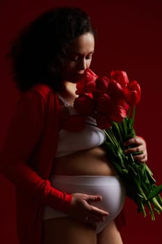 Pregnant woman, expectant mother sniffing a bouquet of red tulips, gently stroking her belly, on isolated background