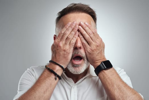 If I ignore the problem, will it go away. Studio shot of a mature man covering his eyes and looking shocked against a grey background.