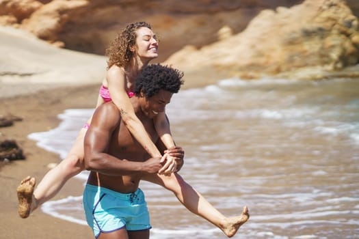 Happy black man giving piggyback ride to girlfriend on beach
