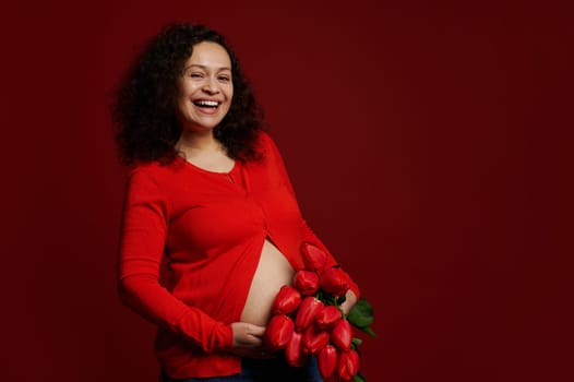 Authentic pregnant woman with big belly, expectant mother, holding a bouquet of red tulips, isolated on red background