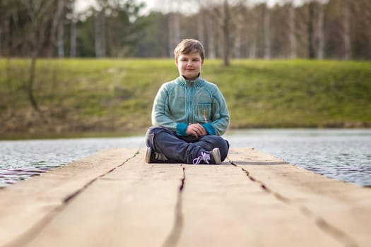 A boy is sitting on a bridge in a green park. The path is a bridge over the lake.