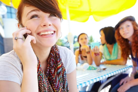 Its great to hear from you. An adolescent girl smiling while she speaks on her cellphone with her friends in the background.