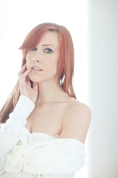 I am desire. Portrait of one beautiful young redhead woman posing seductively in white jersey off shoulder at home. Alluring female feeling flirty, sensual and sexy while teasing with boob cleavage.
