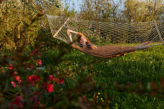 a happy woman in a long orange dress is relaxing in nature lying in a mesh hammock enjoying the peace and tranquility in the country surrounded by green foliage