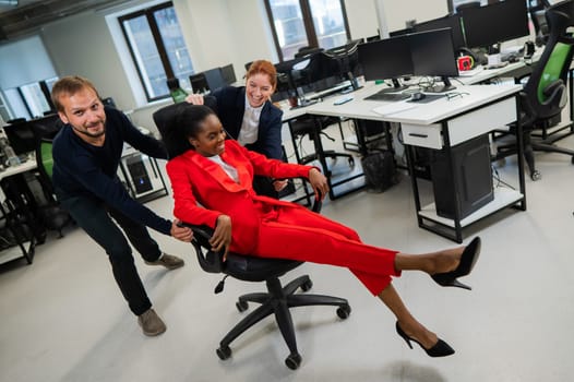Caucasian red-haired woman, bearded caucasian man rolled African American young woman on office chair. Colleagues have fun at work.