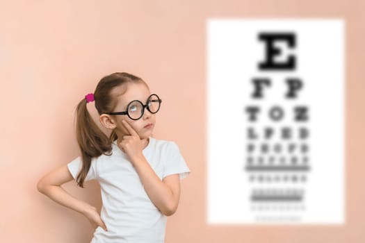 Thoughtful smart girl of elementary school age in diopter glasses against the background of a blurred snellen table for checking visual acuity