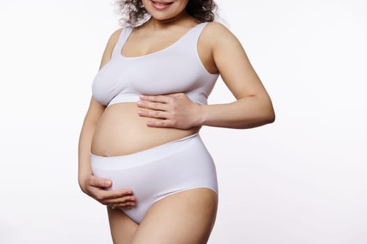 Midsection pregnant woman, expectant mother putting hands on her belly, posing in white underwear on isolated background