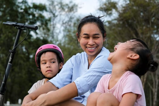 Mother and children sit and rest after riding scooters in the park. Mother and daughters spend their free time riding scooters outdoors together. Happy Loving Family.