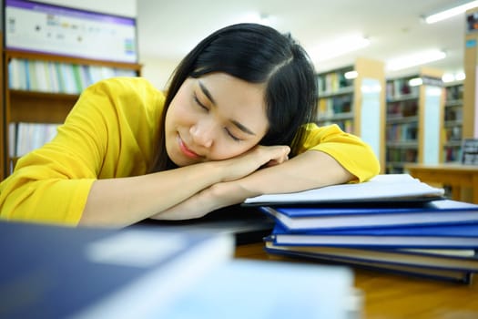 Tired female student sleeping on pile of books in the library with blurred bookshelf on background