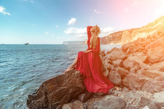 Red dress sea woman. A blonde with flowing hair in a long red dress sits on a rock near the sea. The concept of trips, a photo shoot at the sea.
