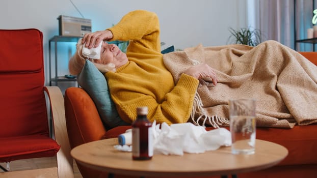 Sick ill mature woman suffering from cold, allergy lying on home sofa sneezes wipes snot into napkin