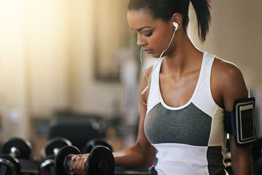 Fitness, music and dumbbell with a woman in the gym, streaming audio through earphones while training. Exercise, workout and weightlifting with a female bodybuilder in a sports club for health