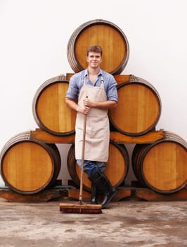 Portrait, broom and oak barrels with a man cleaning the wine cellar of a beverage distillery. Confident, apron and a male cleaner in a winery for the production, fermentation or storage of alcohol