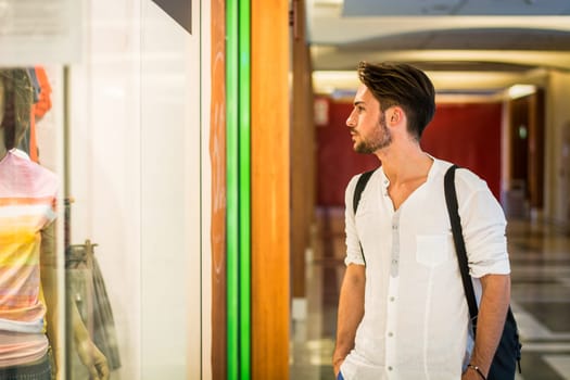 Handsome Young Man in White Shirt and Backpack Looking at Displayed Fashion Items in Glass Window Boutique at the Street Side.