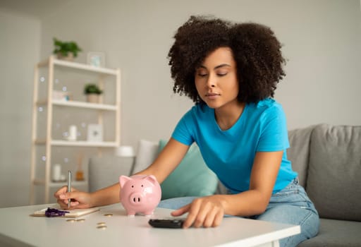 Concentrated millennial curly black lady counting income, tax and mortgage at table with piggy bank