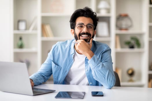 Dreamy indian freelancer man sitting at desk in home office