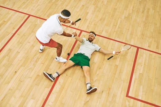 If squash was easy theyd call it tennis. High angle shot of a young man helping his friend up while playing a fame of squash.