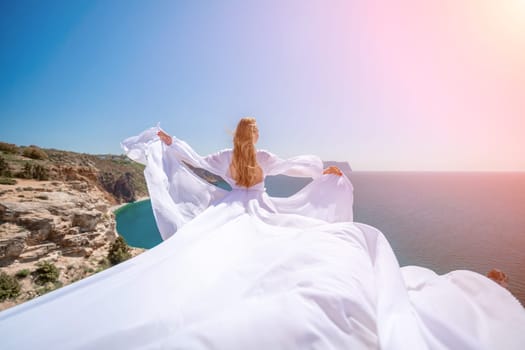 woman sea white dress. Blonde with long hair on a sunny seashore in a white flowing dress, rear view, silk fabric waving in the wind. Against the backdrop of the blue sky and mountains on the seashore