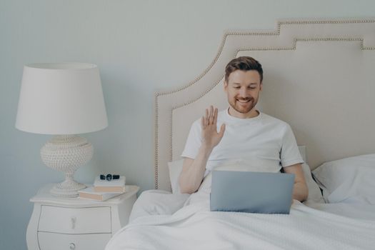 Relaxed young man with stubble using laptop laptop while sitting in bed