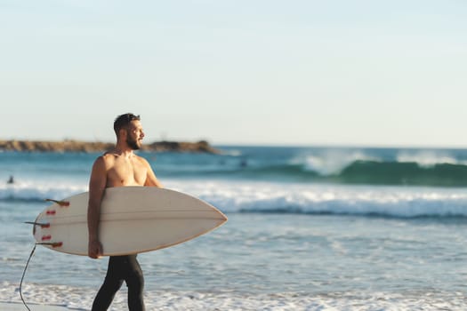 A smiling athletic man surfer with naked torso walking on the seashore