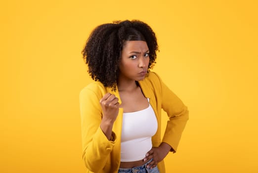Displeased angry black young woman clenching fist, looking with aggression at camera over yellow studio background