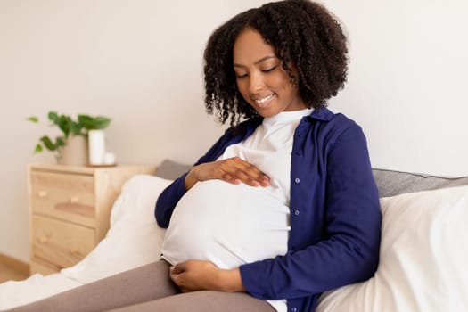 Cheerful curly millennial black woman in domestic clothes with big belly enjoys pregnancy