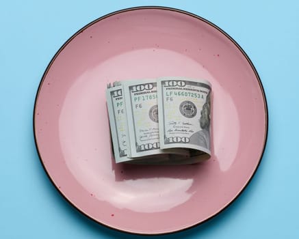 Paper american dollar bills in a ceramic pink plate on a blue background, top view