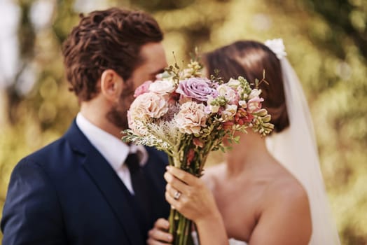 Flowers, wedding and a married couple behind a bouquet together after a ceremony of tradition outdoor. Love, marriage or commitment with a man and woman outside as husband and wife in matrimony