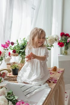 A little blonde girl on a kitchen countertop decorated with peonies. Spring atmosphere.