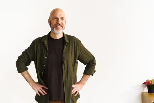 Smiling mid-aged bald man with silver beard standing confidently in front of white wall. His positive posture and expression radiate happiness and joy, showcasing contentment and cheerful demeanor.