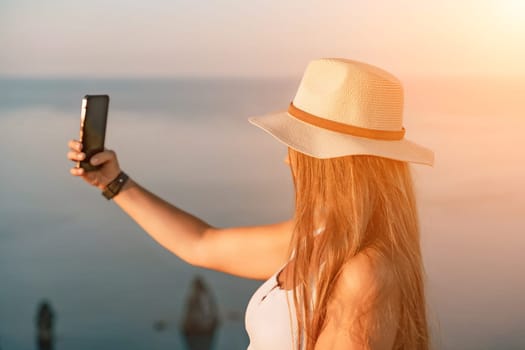 Selfie woman in a hat, white tank top, and shorts captures a selfie shot with her mobile phone against the backdrop of a serene beach and blue sea.