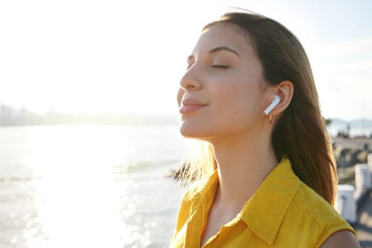 Young woman breathing fresh air with closed eyes listening relaxing music with wireless earphones on city bay at sunset