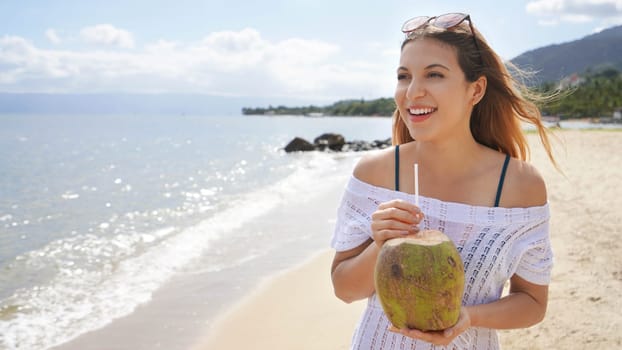 Beautiful young woman holding a fresh coconut enjoying on tropical beach. Girl relax on the beach in her travel holiday holding a green coconut fruit to drink refreshing healthy fresh fruit water.