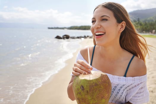 Summer vacation on the beach. Happy relaxed young female tourist drinks green coconut water through a straw and smiling with breeze between hair on her holidays.