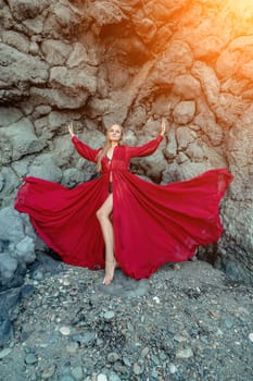 Red dress rocks woman. A blonde with flowing hair in a long flowing red dress stands near a rock of volcanic origin. Travel concept, photo session at sea