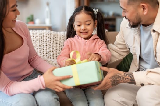 Time For Gifts. Cheerful Japanese Family Celebrating Baby Daughter's Birthday, Giving Wrapped Gift Box To Cute Child Sitting On Sofa At Home. Holiday Celebration Concept. Cropped, Selective Focus