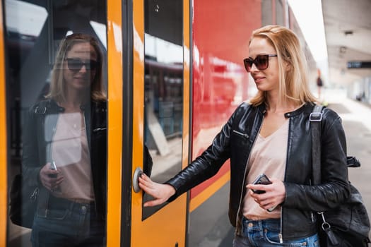 Young blond woman in jeans, shirt and leather jacket wearing bag and sunglass, presses door button of modern speed train to embark on train station platform. Travel and transportation.