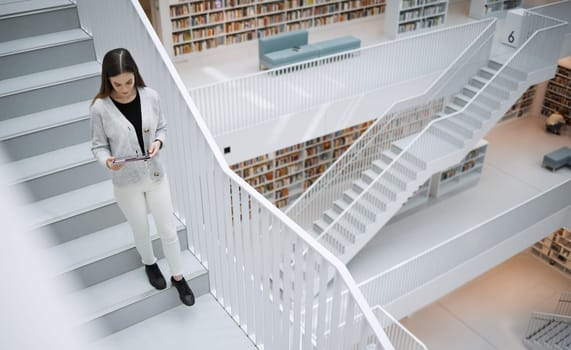Top view, stairs and woman in library, tablet or online research for university, reading or education. Female, girl or student with device, bookshelves or search internet for knowledge or information.