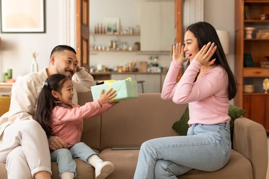 Time For Presents. Cheerful Asian Father And Baby Daughter Congratulating Mother Giving Gift Box To Her Sitting On Couch At Home. Family Celebrating Mom's Birthday Together