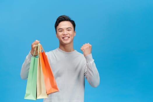 Smiling Asian man holding shopping bags raised fist up shout yeah isolated on blue background