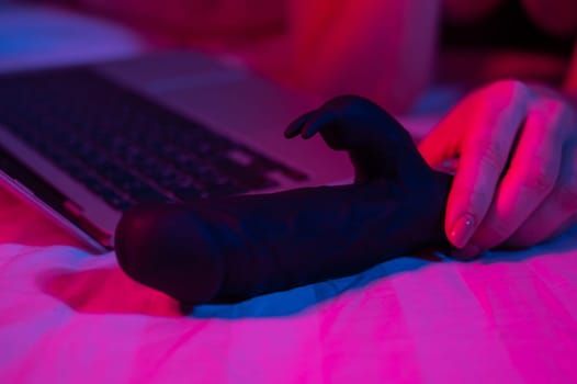 A woman lies in bed with a black dildo and looks at a laptop. Girl using sex toy in blue-red light.