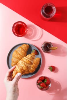 A woman's hand holds a kraussant over a plate with fresh puff croissants, berry juice, jam and fresh berries on a pink and red background.