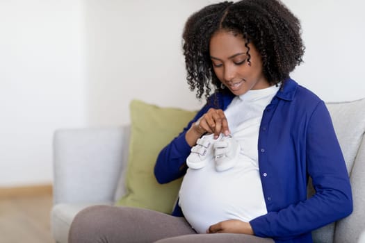 Glad curly young black woman in domestic clothes with big belly enjoy pregnancy, hold small shoes