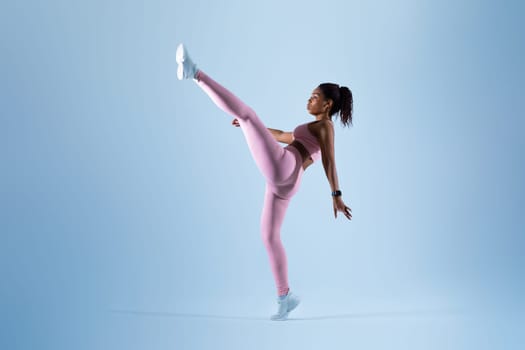 Motivated black lady doing karate kick move, raising leg and showing perfect stretching, blue background, full length
