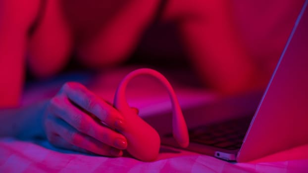 A woman is lying in bed holding a curved vibrator and looking at a laptop. Girl using sex toy in blue-red light.