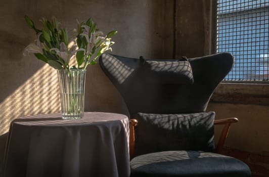 A Wooden Upholstered Chair and the Bouquet of Flowers in a Glass Vase on Table in the Room with Bare Cement Wall. 