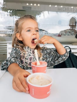Little girl eating ice cream from two cups with a spatula at the table